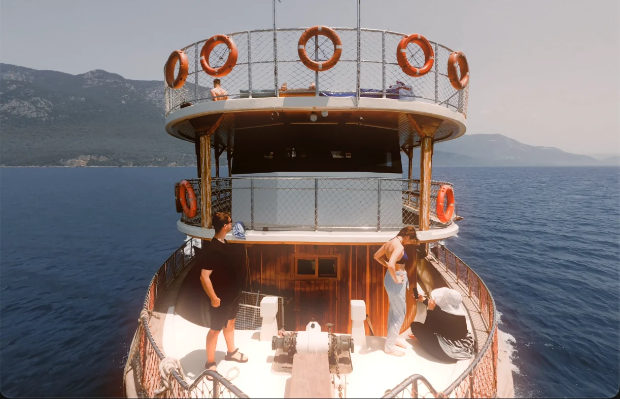 Source frame: a wooden boat with people on deck, sea and hills behind, before grading